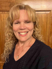 Smiling woman with blonde curly hair in a black top standing before a wooden door