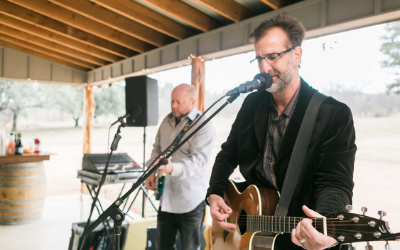 Musician singing and playing acoustic guitar on a covered stage, with a keyboardist behind him