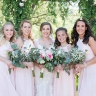 Five women in light pink dresses holding bouquets, standing outdoors beneath trees.