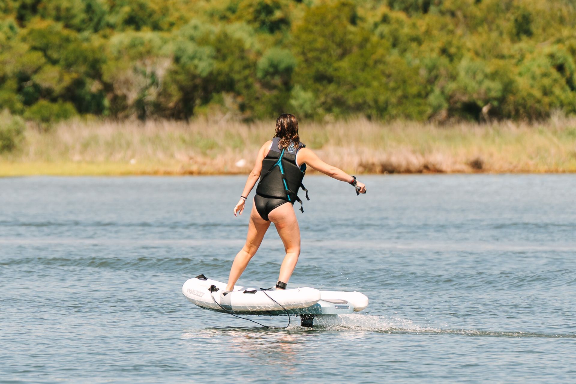 Person eFoiling on calm water near a grassy shoreline with trees in the background