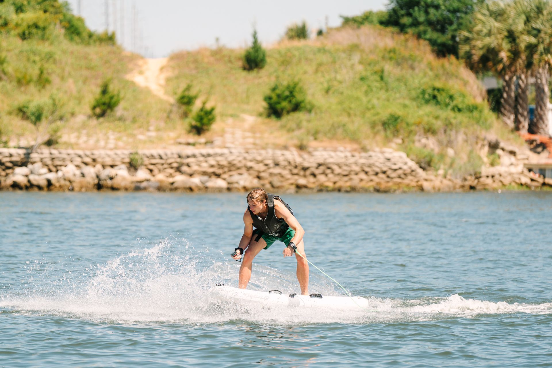 Person eFoiling on a lake, leaning forward and splashing water near a grassy shore.