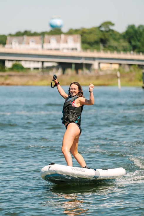 Woman standing on a eFoil board, holding a paddle on a river with a bridge in the background