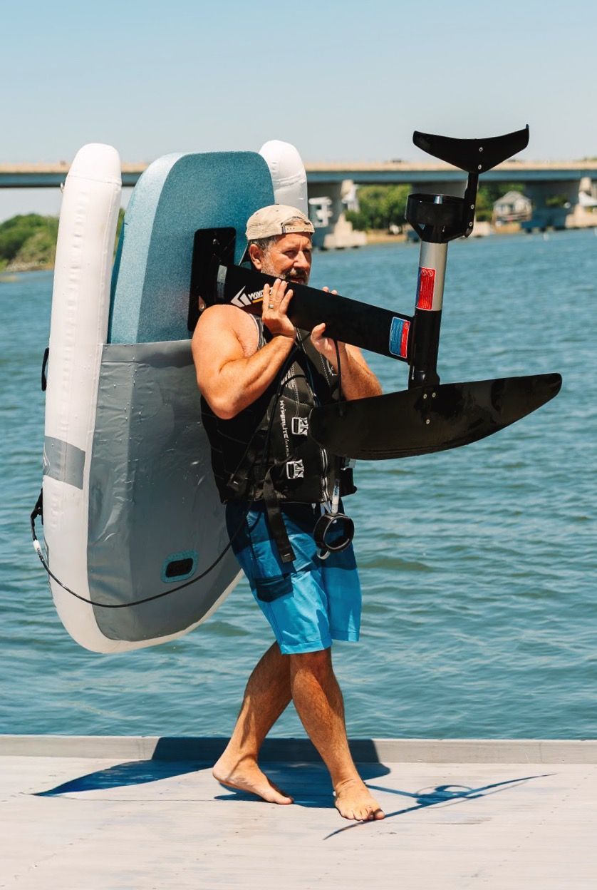 Man carrying a black eFoil board on a dock by the water, with a bridge in the background