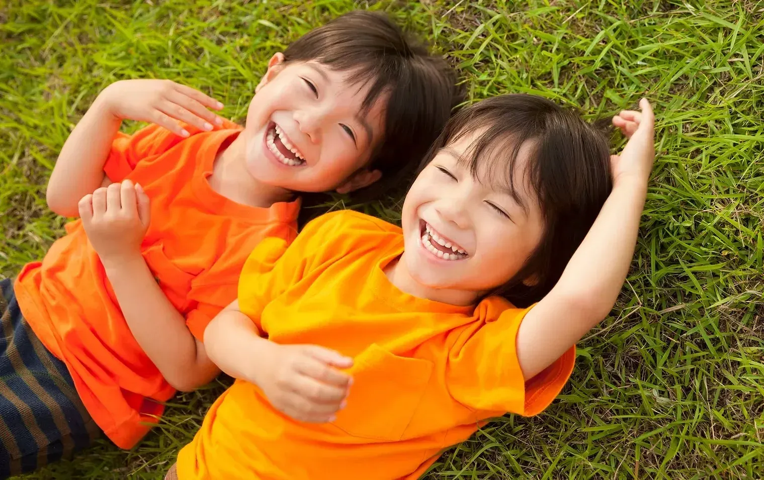 Two children in bright orange shirts laughing and lying on grass