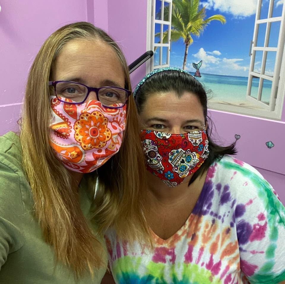 Two masked women posing indoors, with a beach mural background and colorful shirts