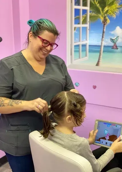 Hairdresser styling a child’s hair in a pink salon, with a beach mural in the background