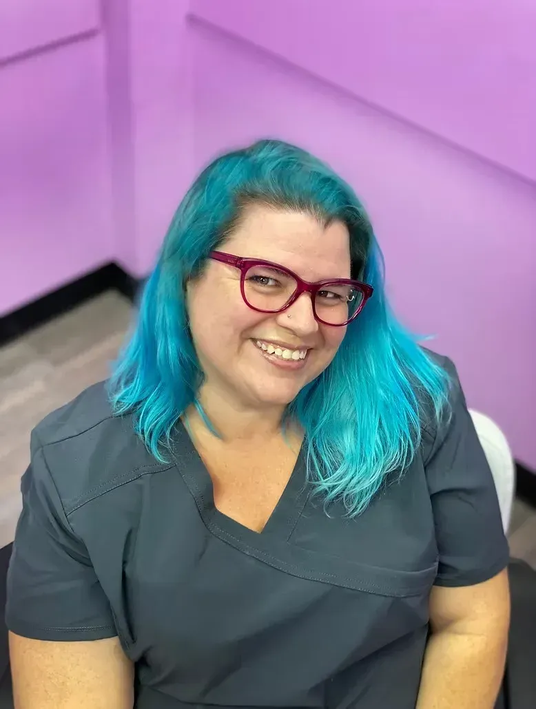 Smiling person with teal hair and magenta glasses sitting against a lavender wall