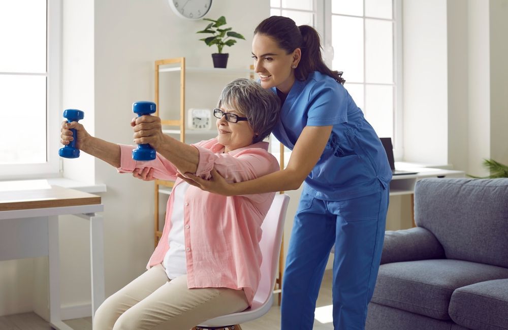 A Caregiver in Blue Scrubs Assists an Individual Sitting in a Chair — Elevate Injury Management in Sandy Bay, TAS