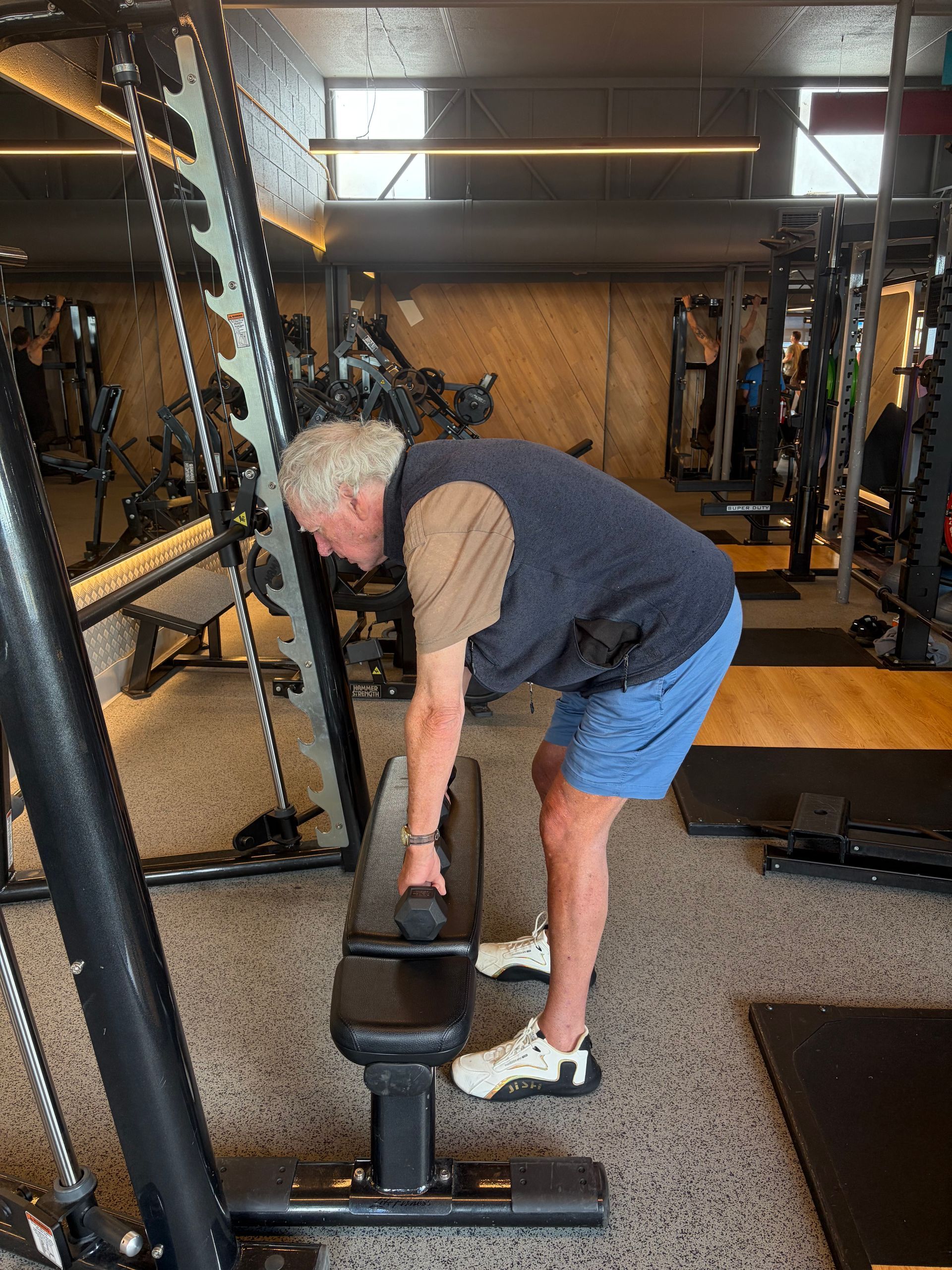 Man bending to lift a dumbbell in a gym with mirrors and weight equipment — Elevate Injury Management in Sandy Bay, TAS