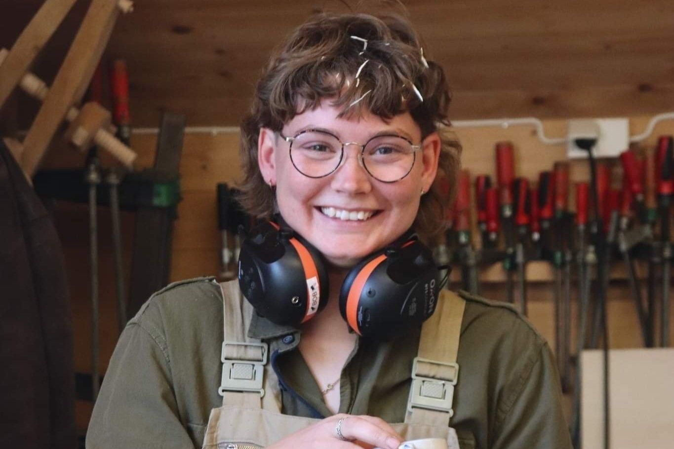 A woman wearing headphones and glasses is smiling in a workshop.