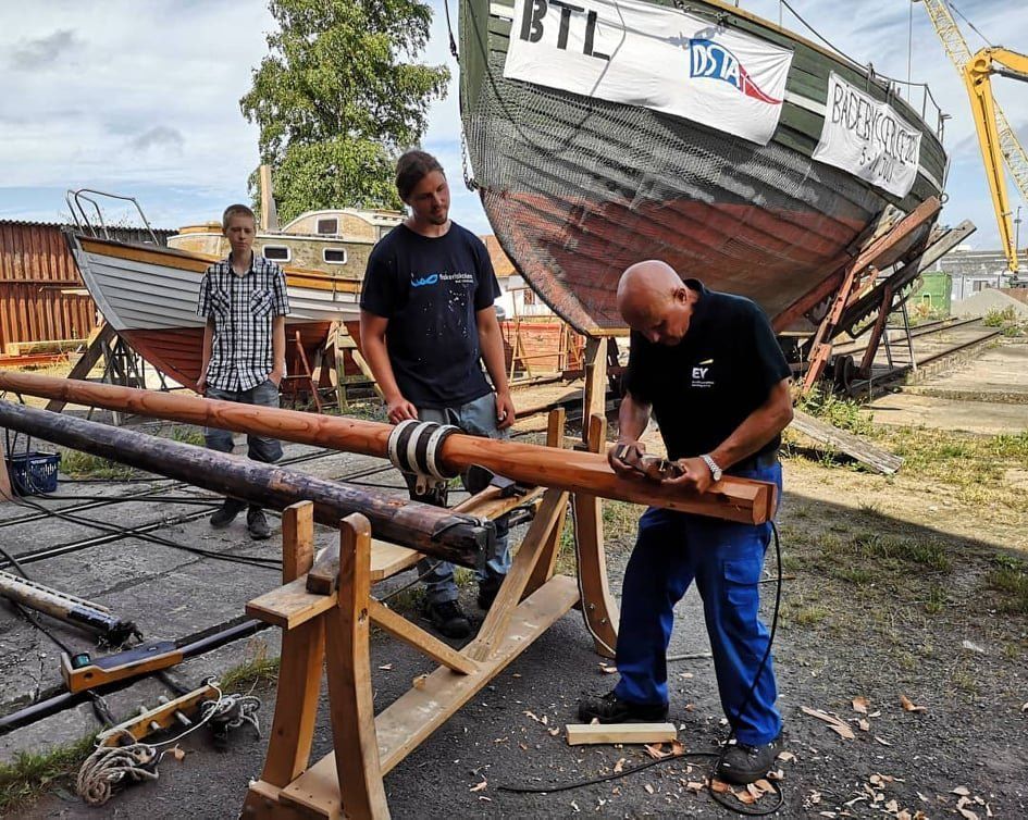 A man is carving a piece of wood in front of a boat.