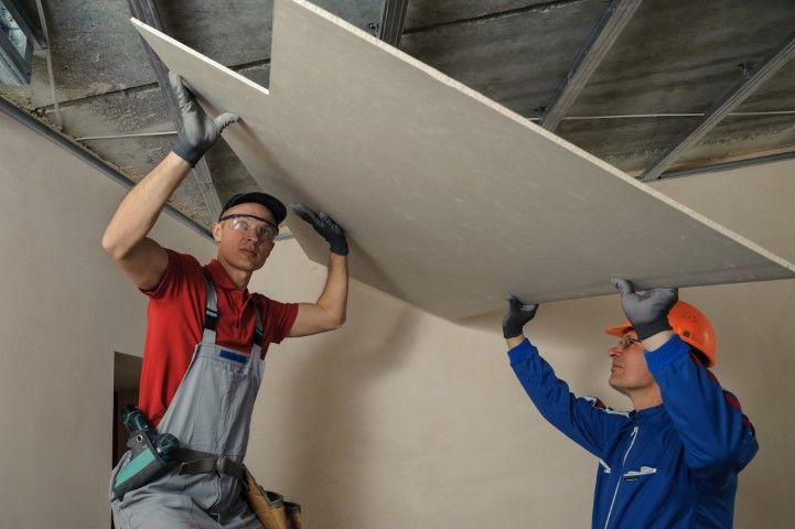 Two workers installing drywall on a ceiling. One wears a red shirt, the other a blue jumpsuit, both wearing gloves.