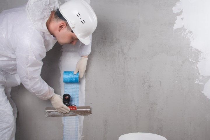 Person in protective suit applying coating to a wall with a roller and trowel.