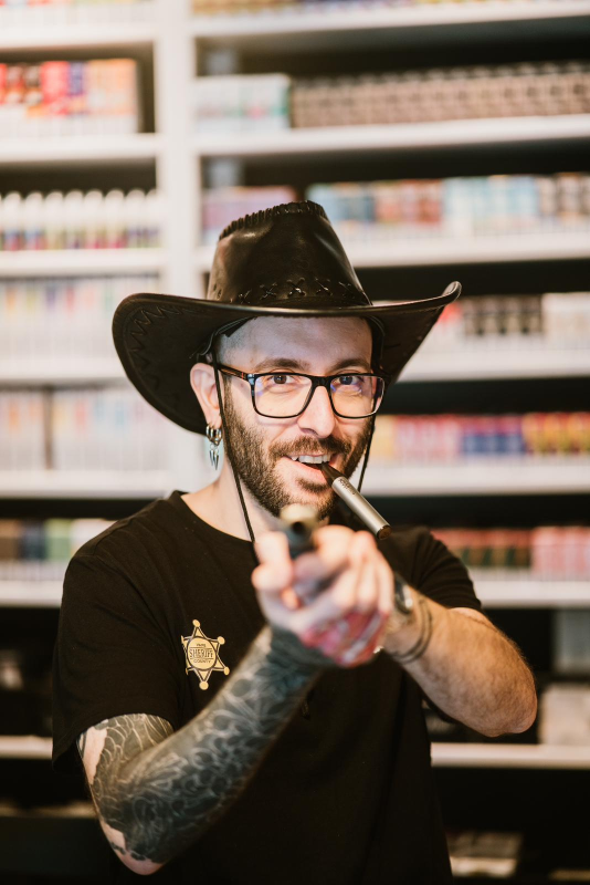 Un uomo con cappello da cowboy e occhiali indica una sigaretta elettronica; sullo sfondo sono esposti scaffali di prodotti.