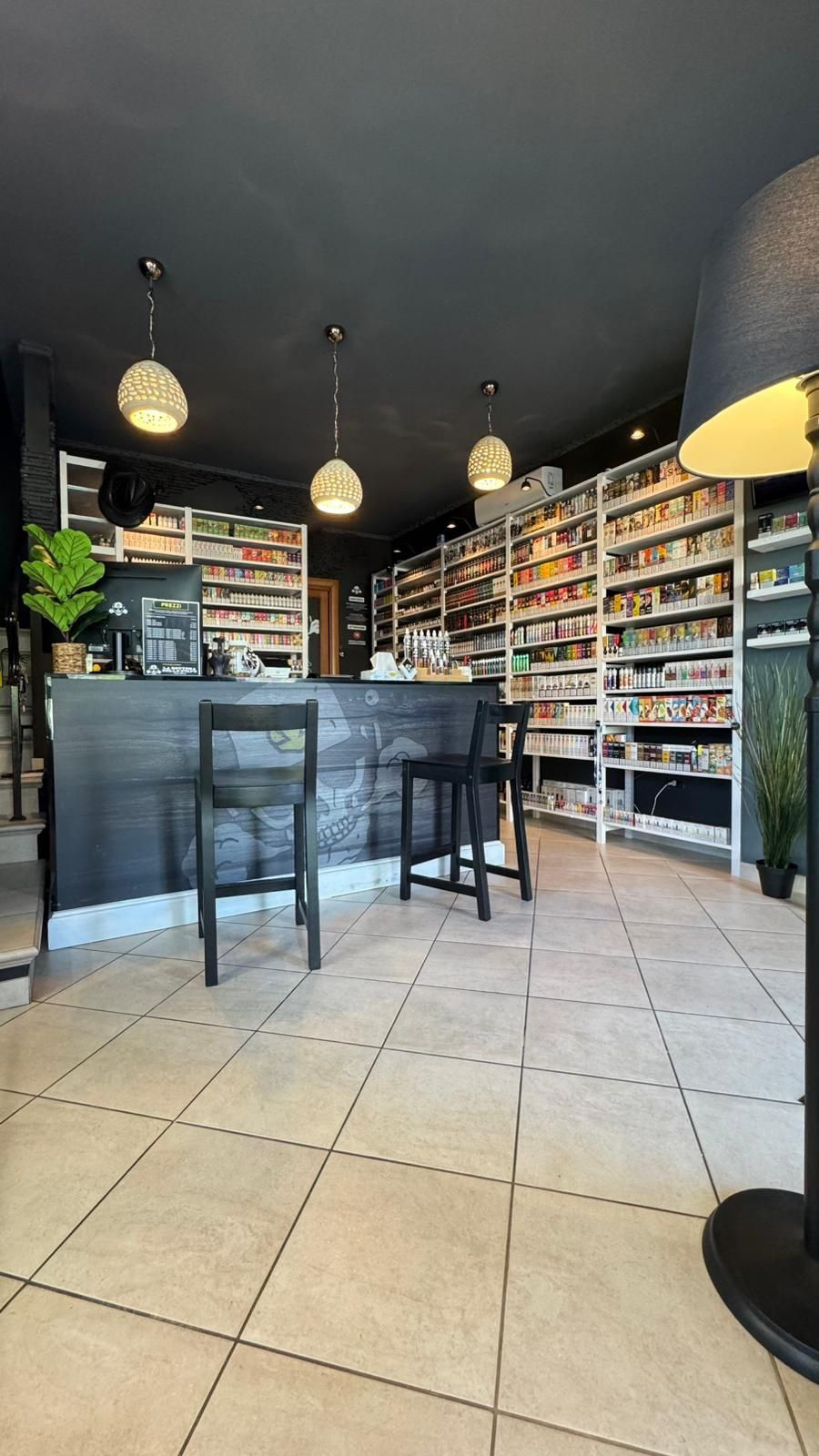 Bar con scaffali pieni di oggetti, due sedie e lampade a sospensione sferiche. Pavimento piastrellato e soffitto nero.