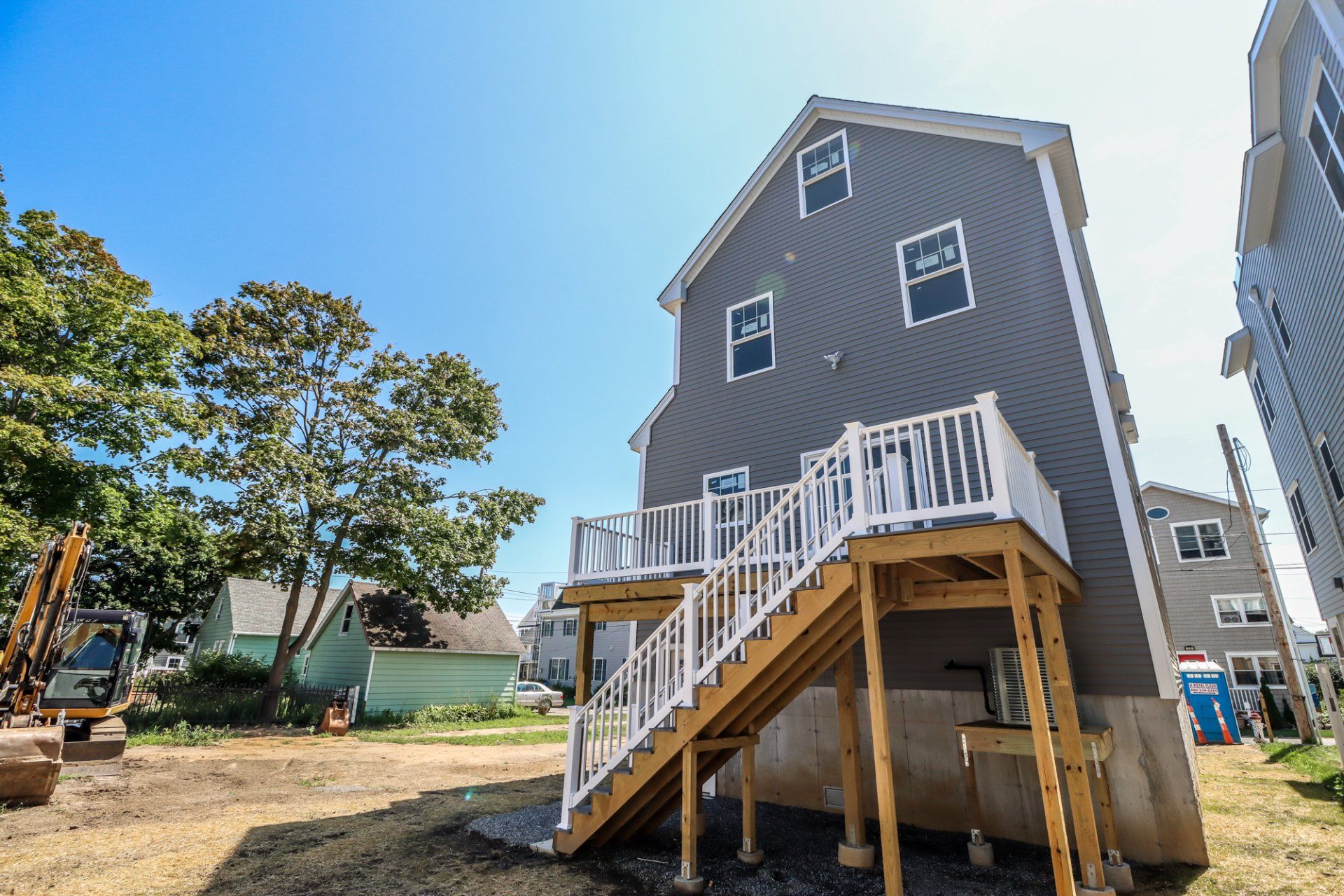 Back of a gray three-story house with a wooden deck and stairs. Construction site with blue sky.