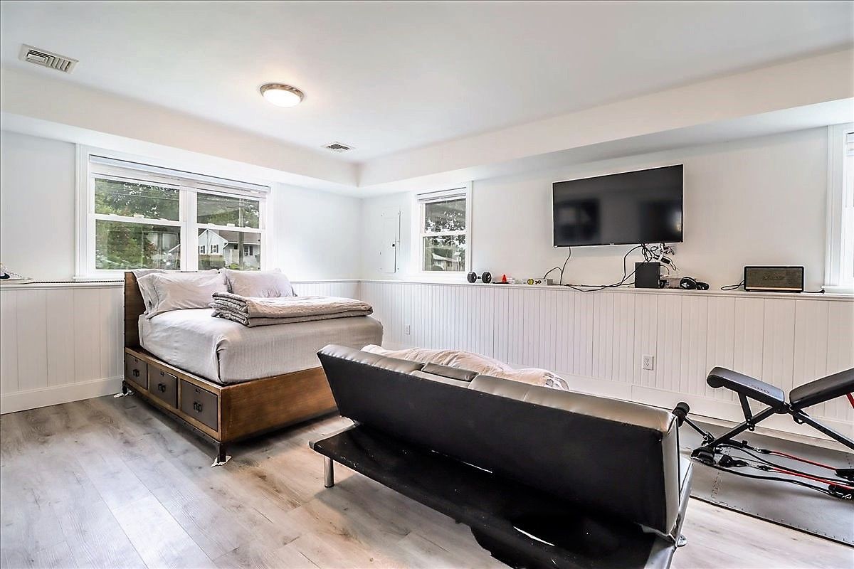 A modern, white-walled room with a bed, futon, TV, and workout equipment near two windows.