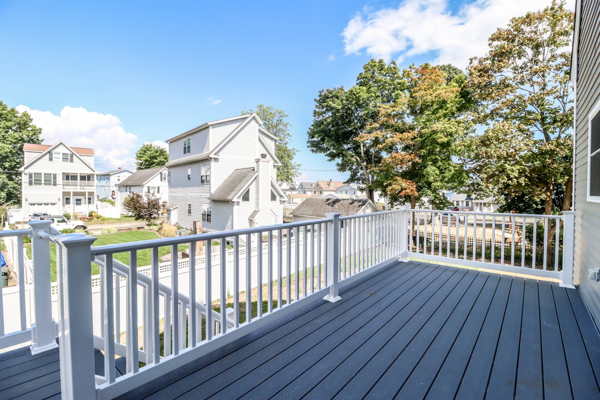 A wooden deck with white railing overlooks neighborhood houses and trees under a blue sky.