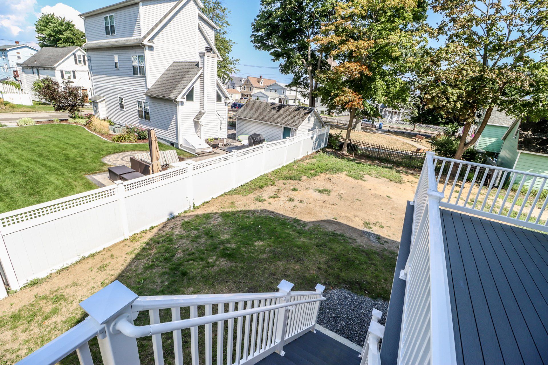 Backyard with white fence, stairs, gray deck, and green lawn. Houses in background.