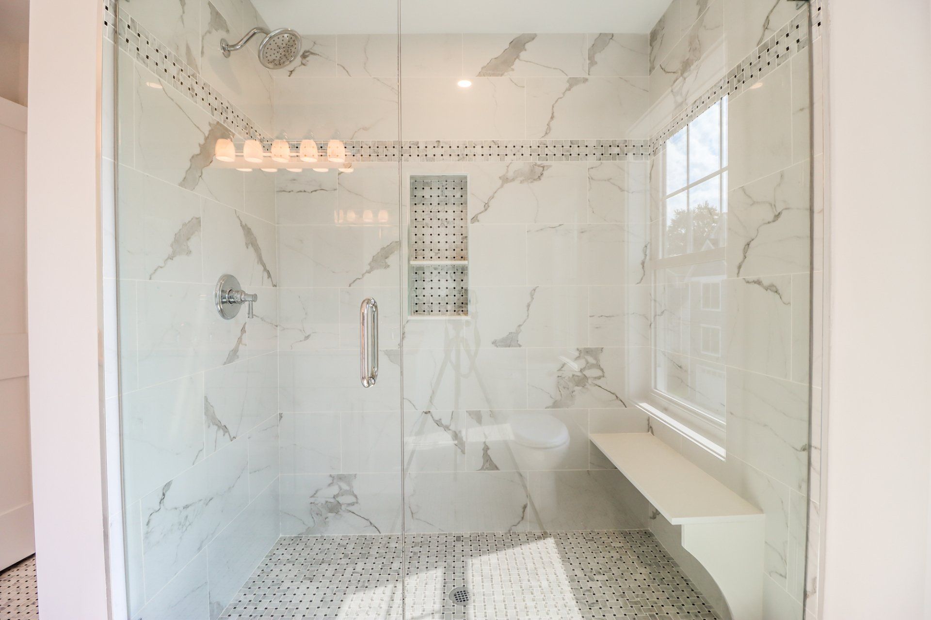 A modern shower with marble-like tiles, a glass door, a built-in bench, and a small window.