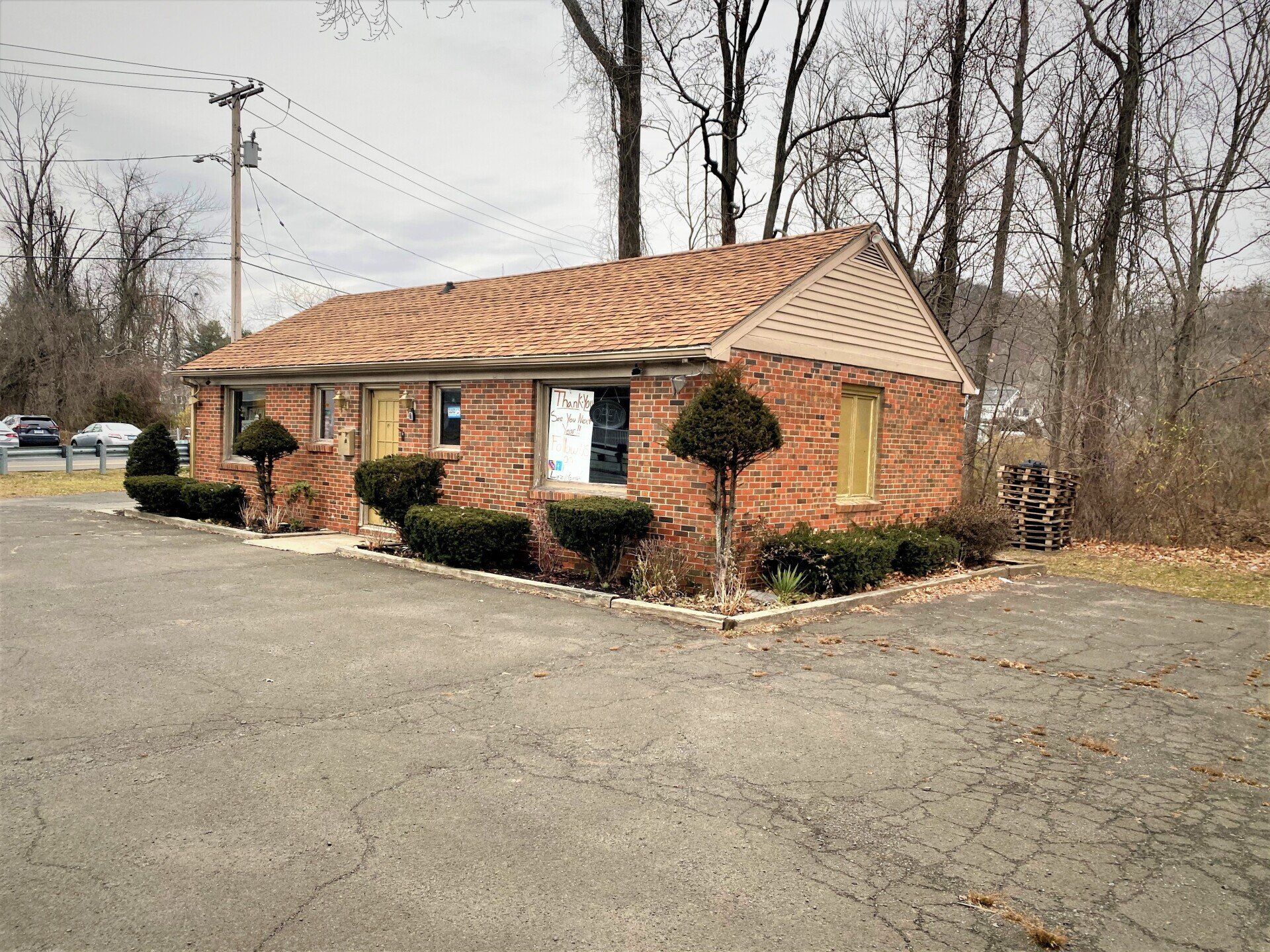 Brick building with a brown roof and yellow window frames, set on asphalt with bare trees behind.