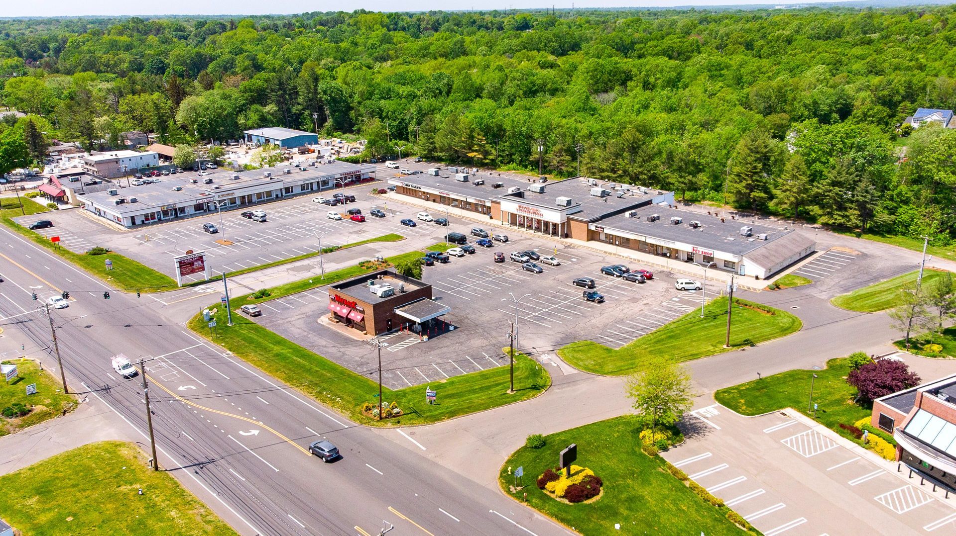 Aerial view of a commercial plaza with parking, surrounded by trees. Includes a red-roofed building, and a road.