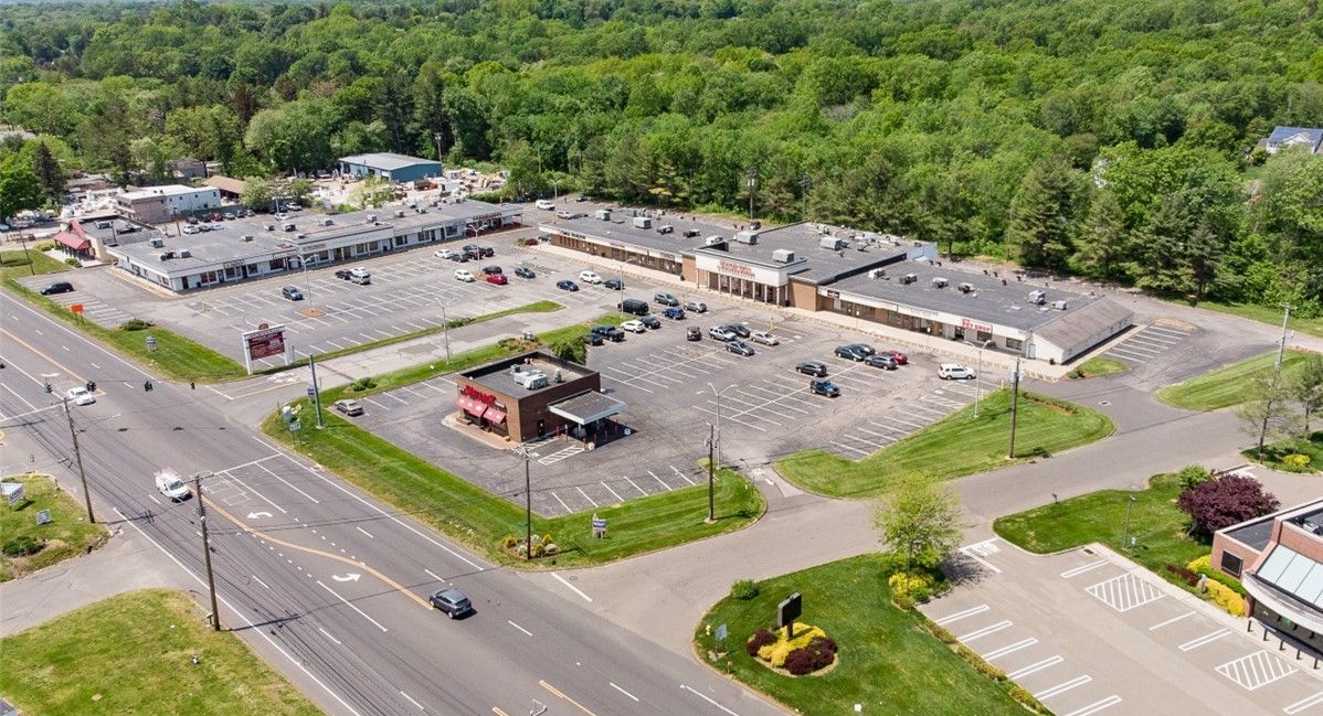 Aerial view of a strip mall with a parking lot next to a busy road, trees in background.