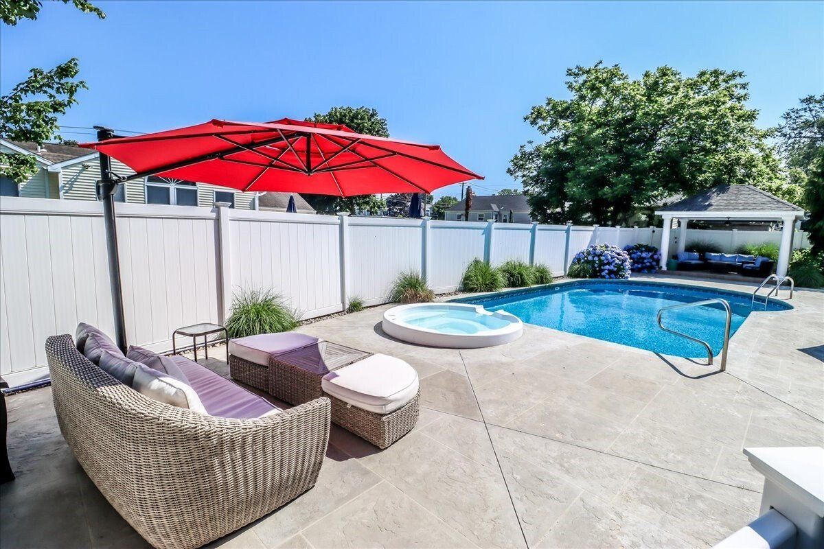 Backyard with pool, hot tub, and patio furniture under a red umbrella on a sunny day.