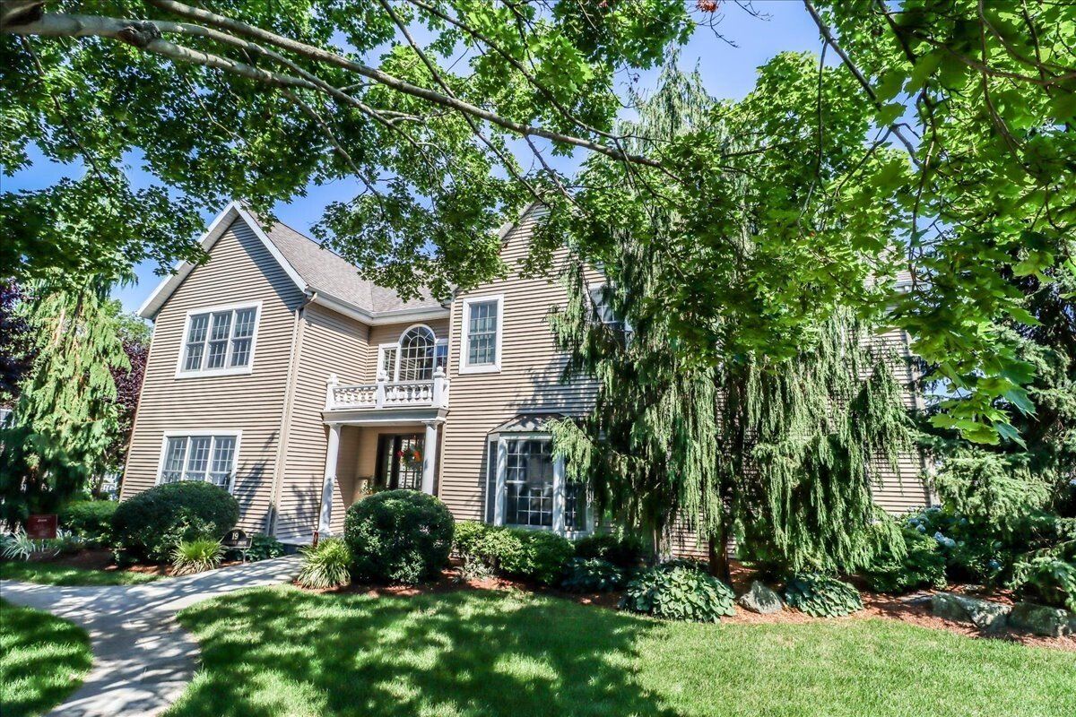 Two-story beige house with a walkway, surrounded by trees and green lawn on a sunny day.