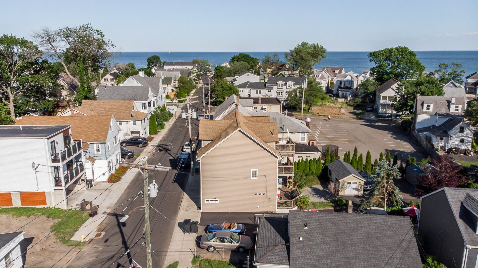 Aerial view of a street lined with houses, leading to a body of water under a blue sky.