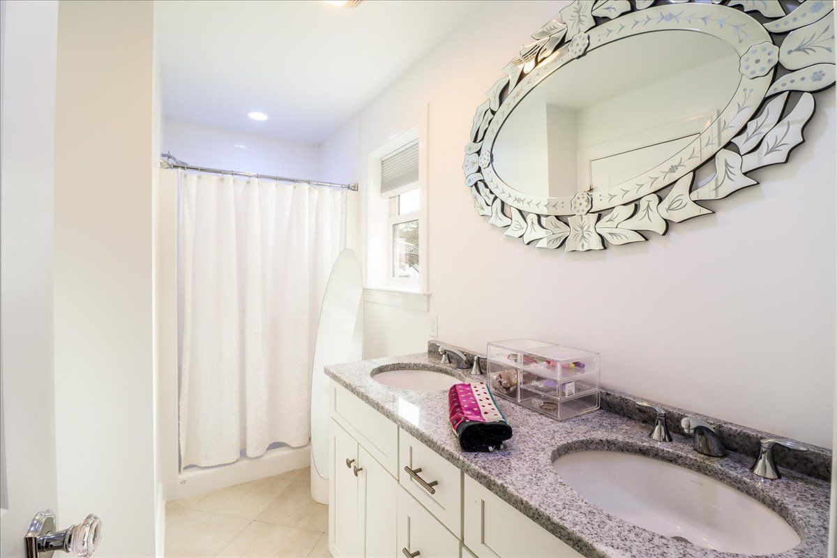 Bathroom with white cabinets, speckled countertop, oval mirror, shower curtain, and two sinks.