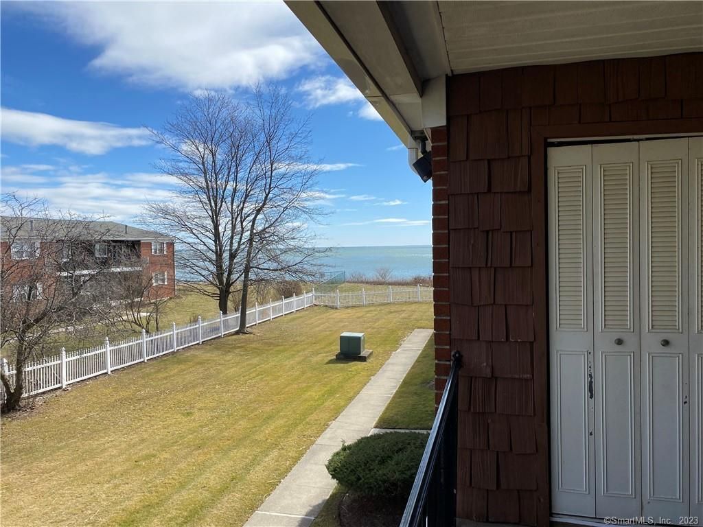 View from balcony overlooking grass, white fence, building, tree, and body of water under a blue sky.