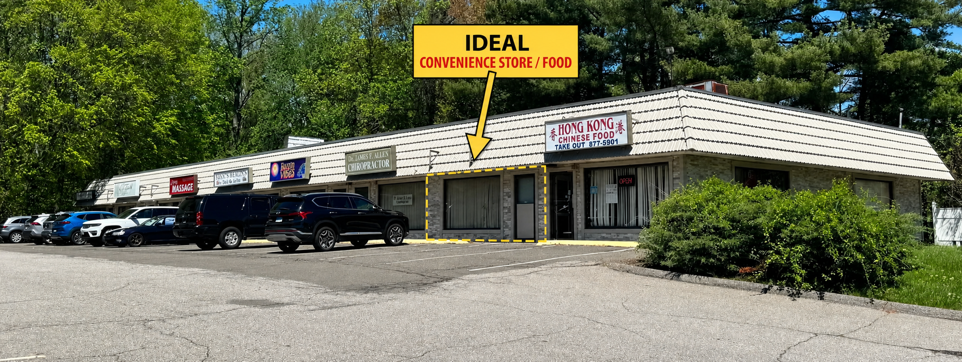 Strip mall storefront with yellow “IDEAL” sign, parked cars, and bushes on a sunny day