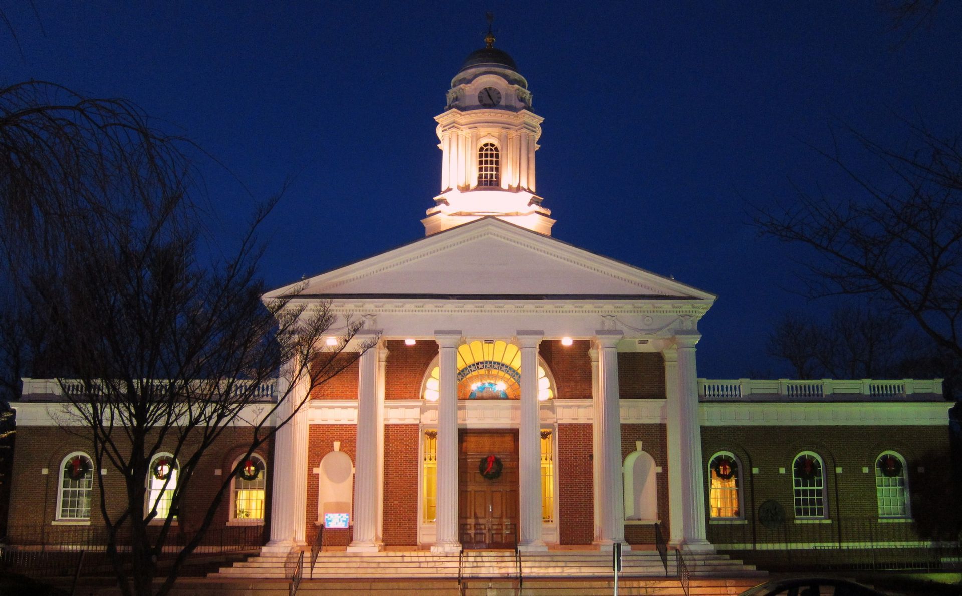Brick building at night with white columns, a tall cupola, and Christmas lights.