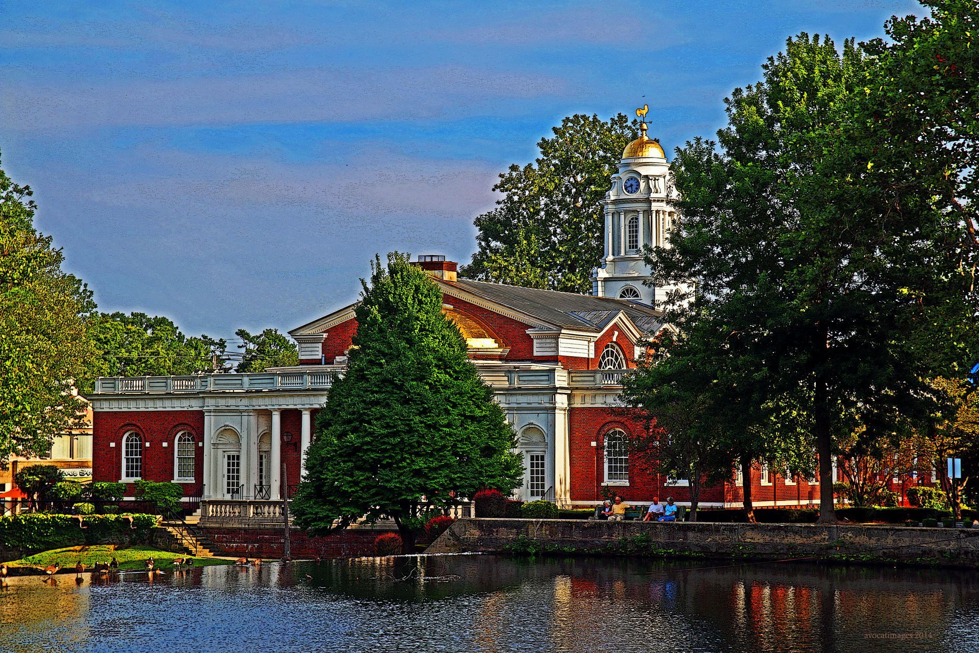 Brick building with columns and gold dome reflected in calm water with trees.
