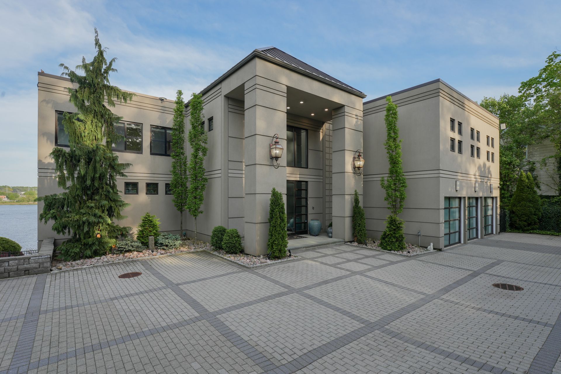 Modern gray house with brick driveway, trees, and lake view.