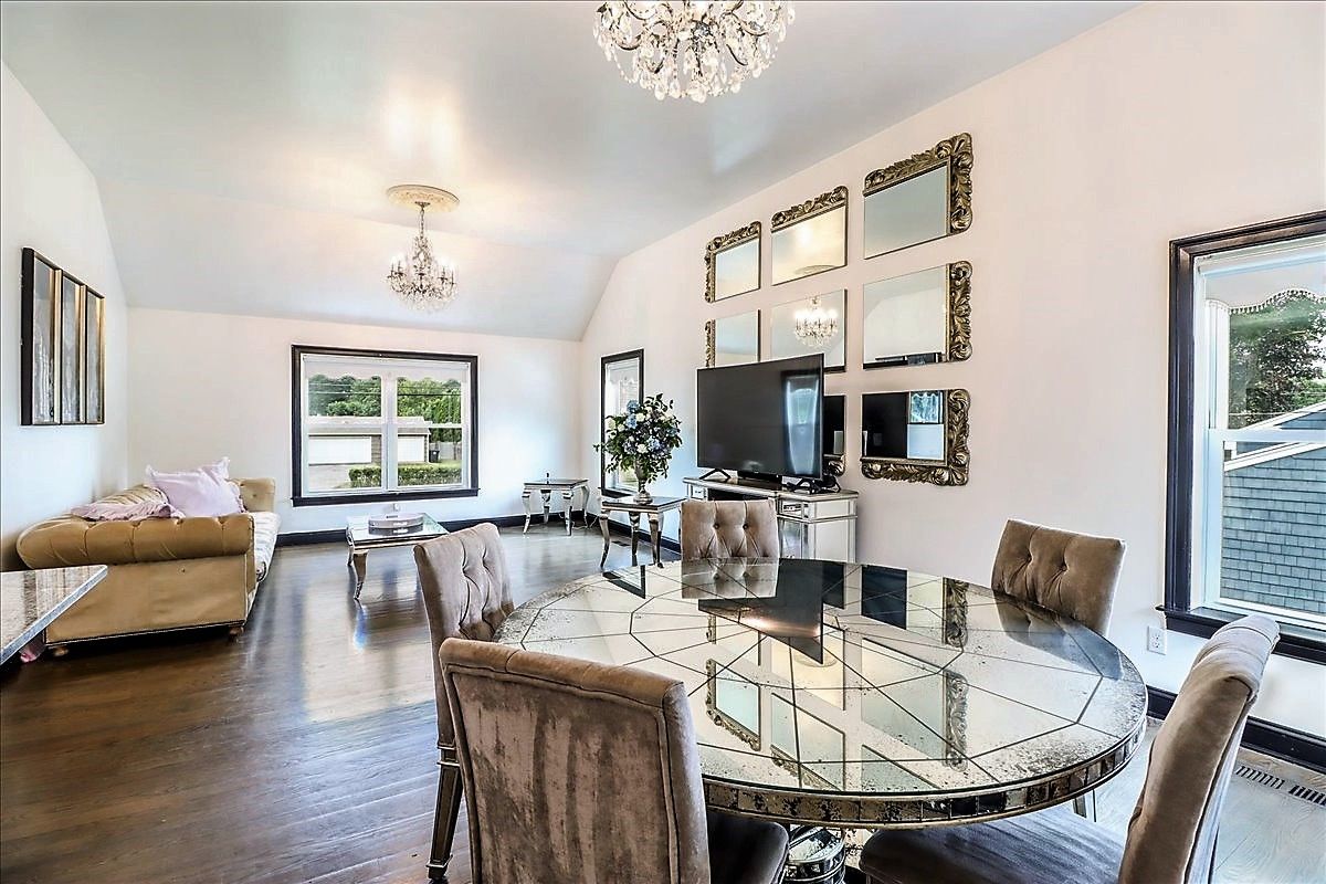 Living room with dining area. Mirrors on wall, round mirrored table, velvet chairs, brown sofa, chandeliers, large windows.