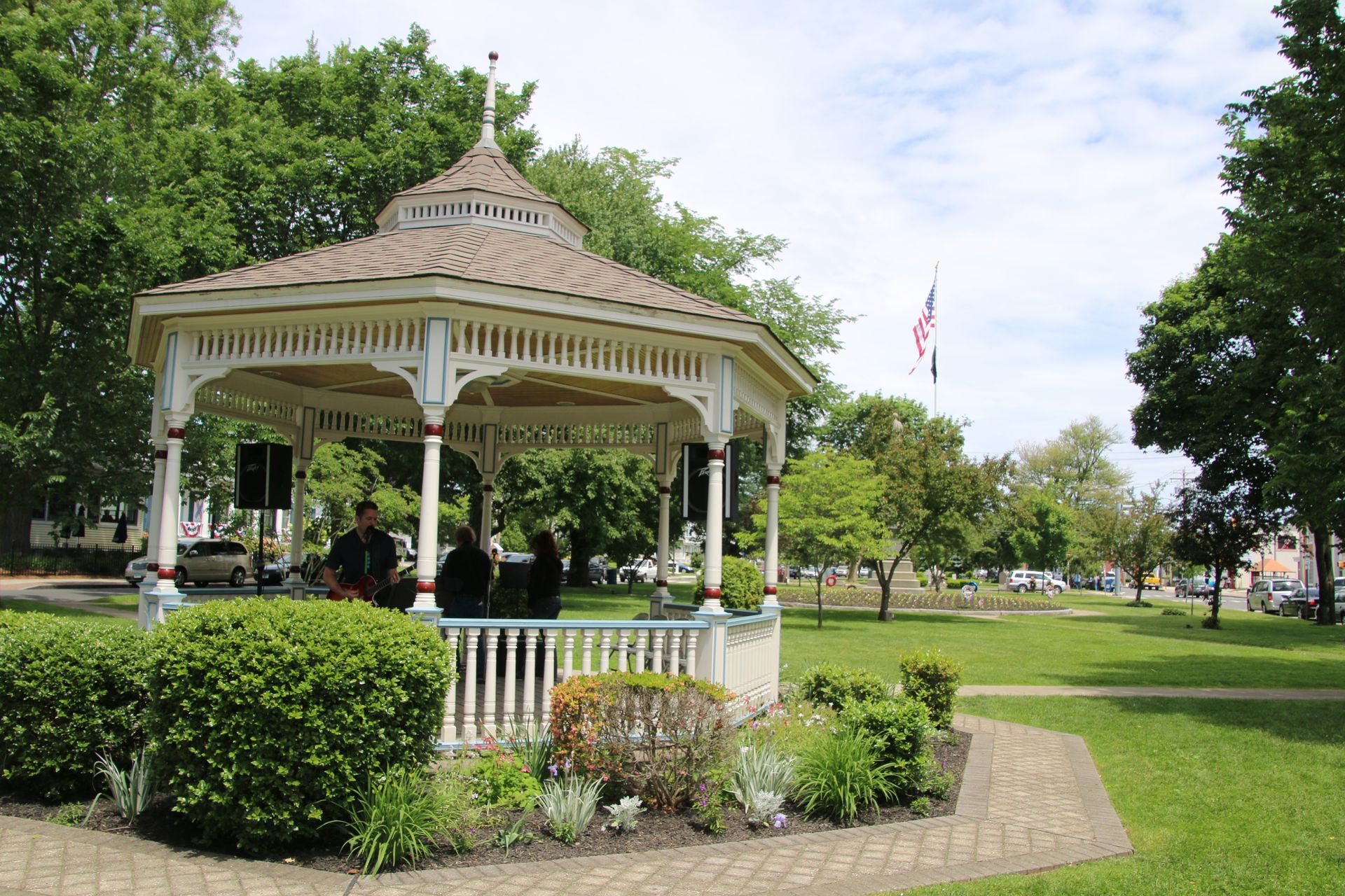 Band playing in a white gazebo in a park, green grass, trees, and American flag.