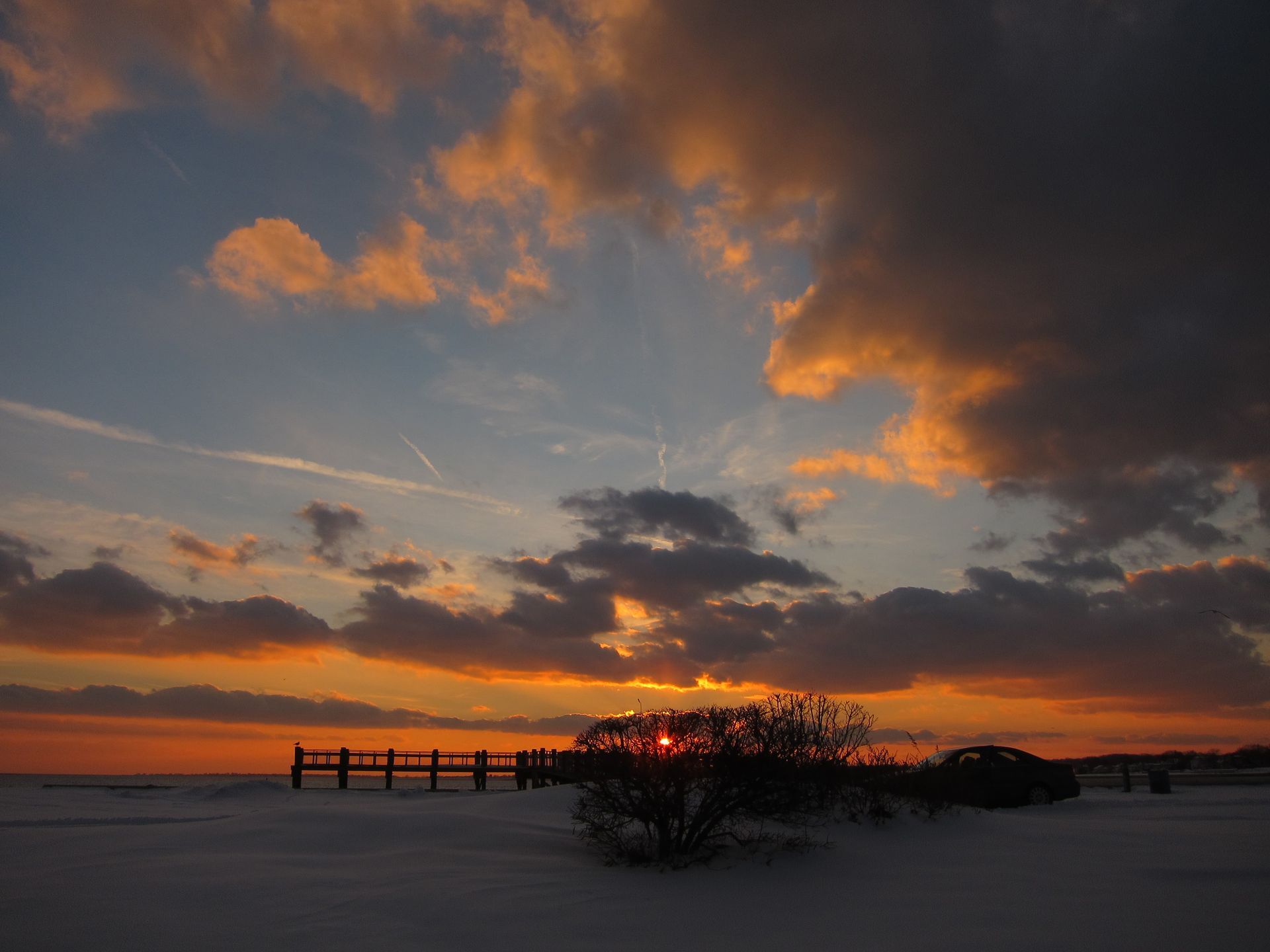 Sunset over snow-covered landscape; orange, blue, and gray clouds; silhouetted trees and pier.