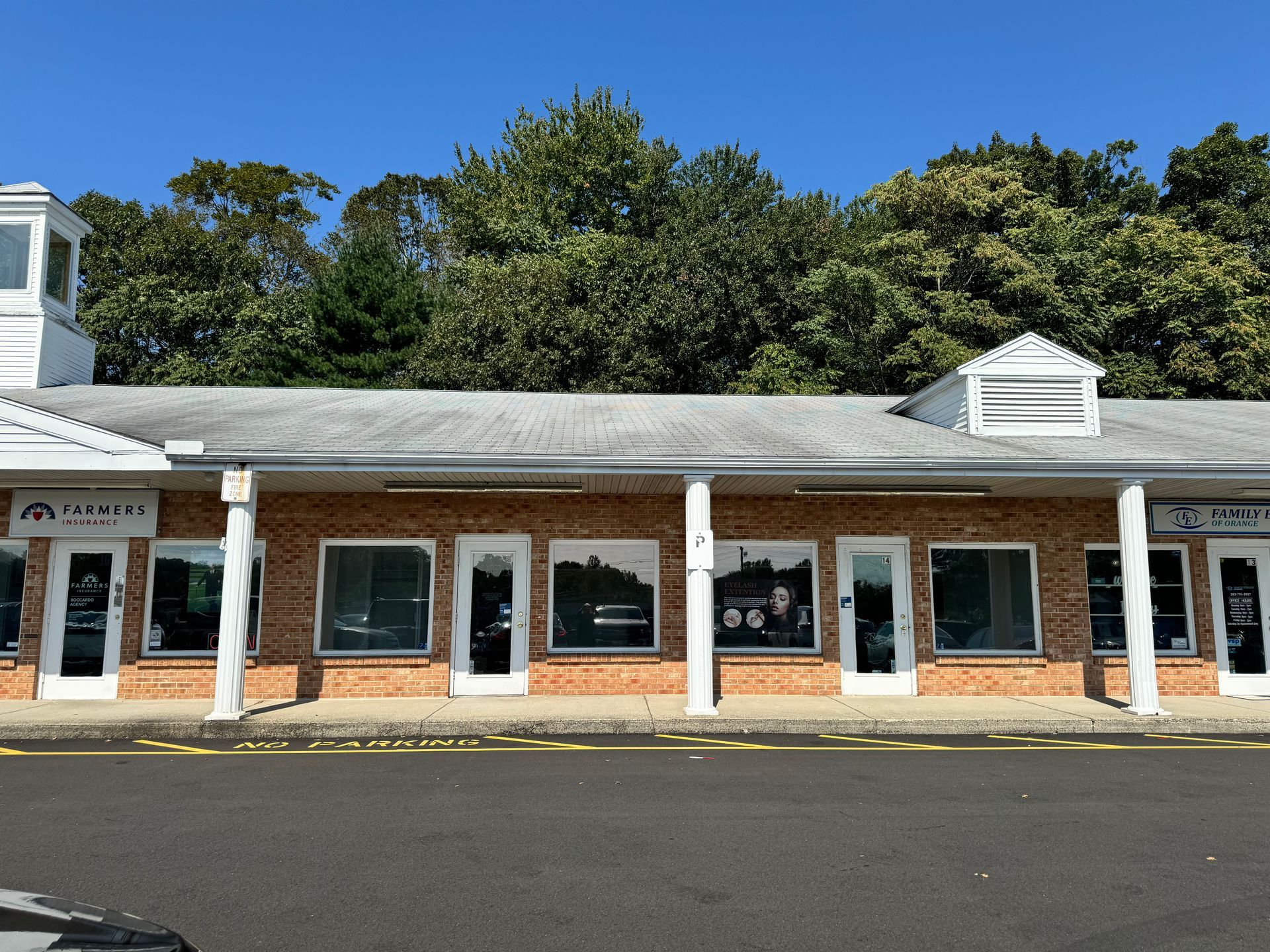 Brick storefront building with white columns and windows under a blue sky, trees in background.