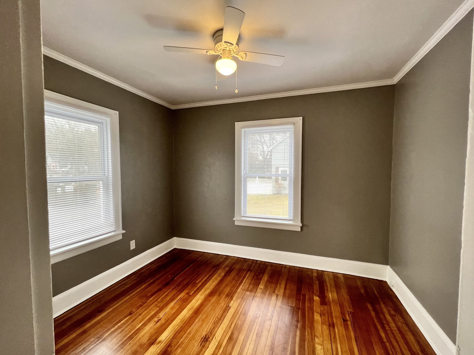Empty room with brown walls, hardwood floors, two windows, and a ceiling fan.