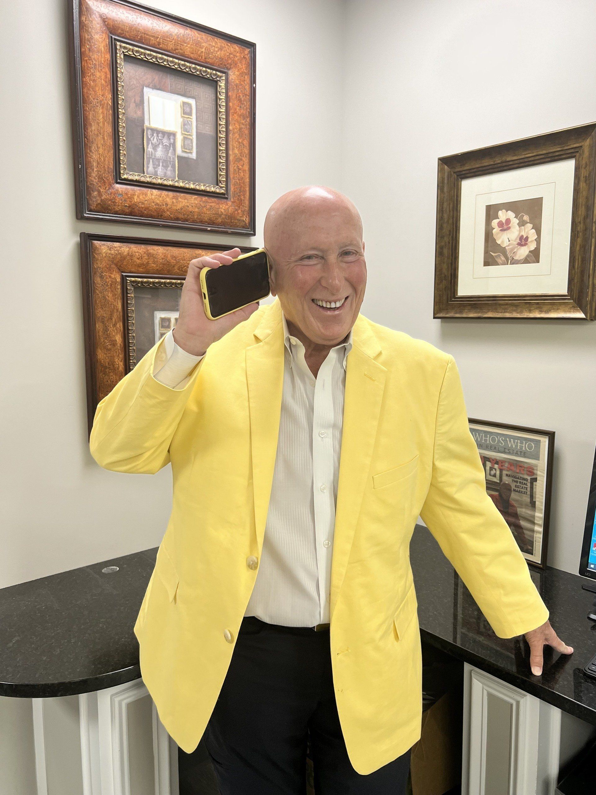 Man in yellow blazer holding a phone near his ear, smiling. Office setting, framed art on wall.