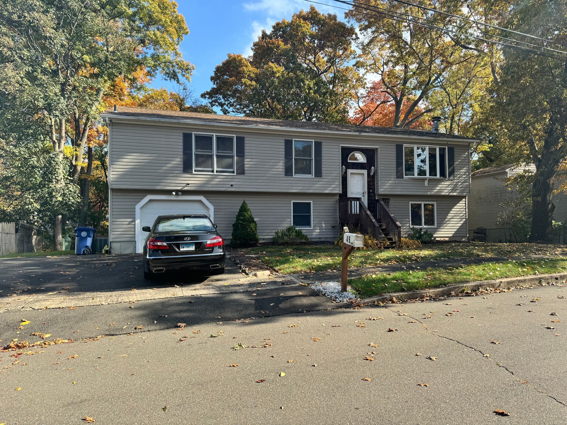 Gray two-story house with a black car in the driveway, surrounded by trees with fall foliage.