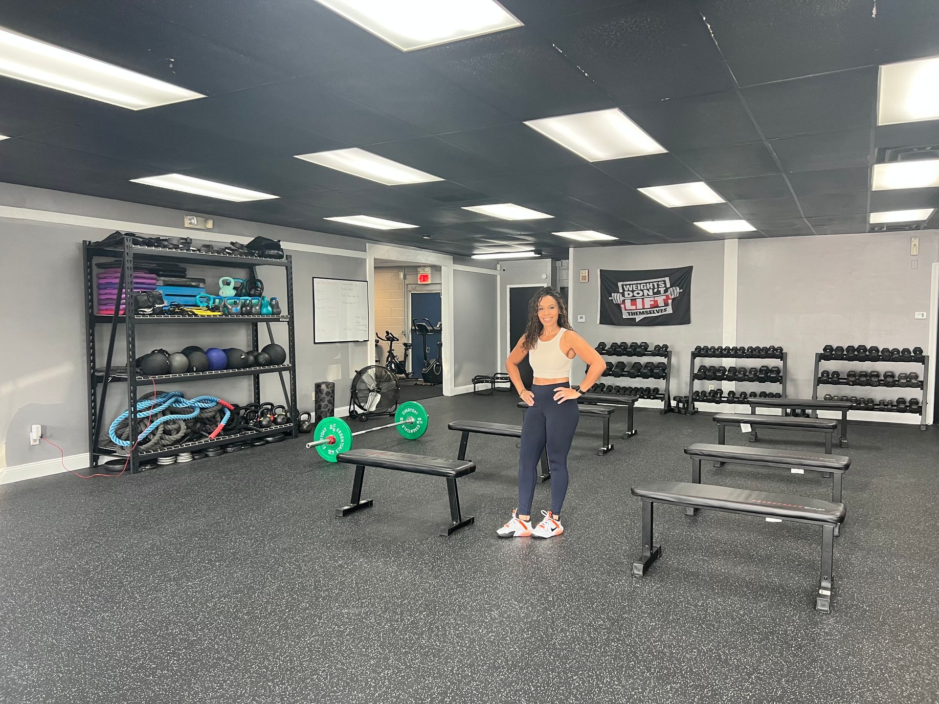 Woman stands in gym, arms on hips, near benches and weights. Gray walls and dark flooring.