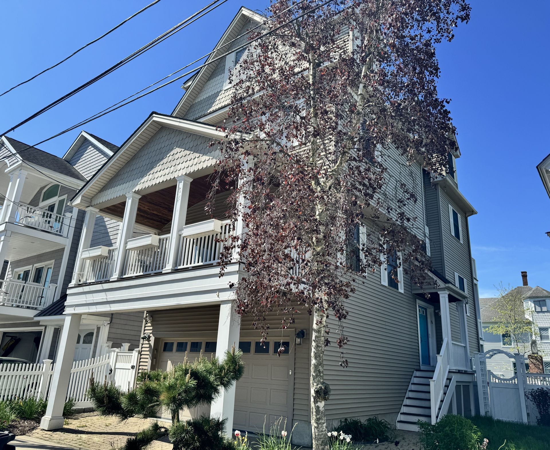 Multi-story house with light siding, a garage, and a porch under a blue sky, with a tree in front.