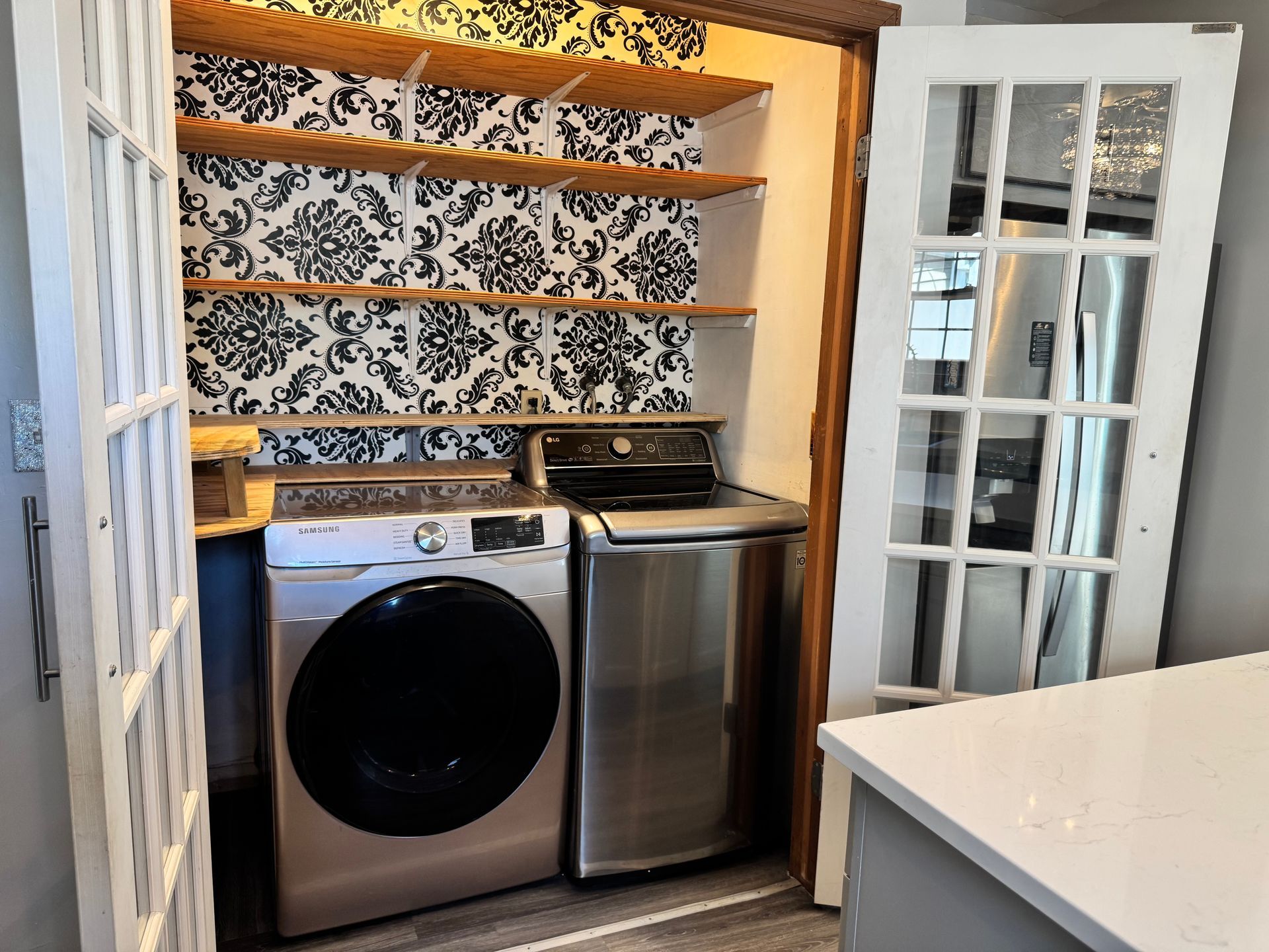 Laundry closet with washer, dryer, shelving, and patterned wallpaper. White doors are open.