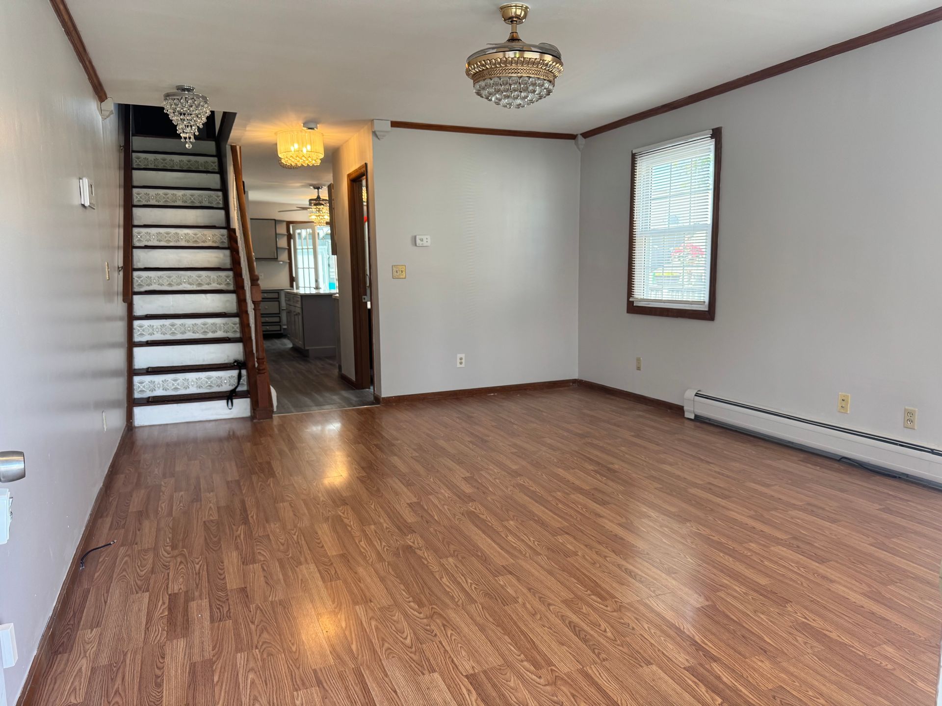 Empty living room with wood-look flooring, staircase, and chandelier lighting. Window with blinds on the right.