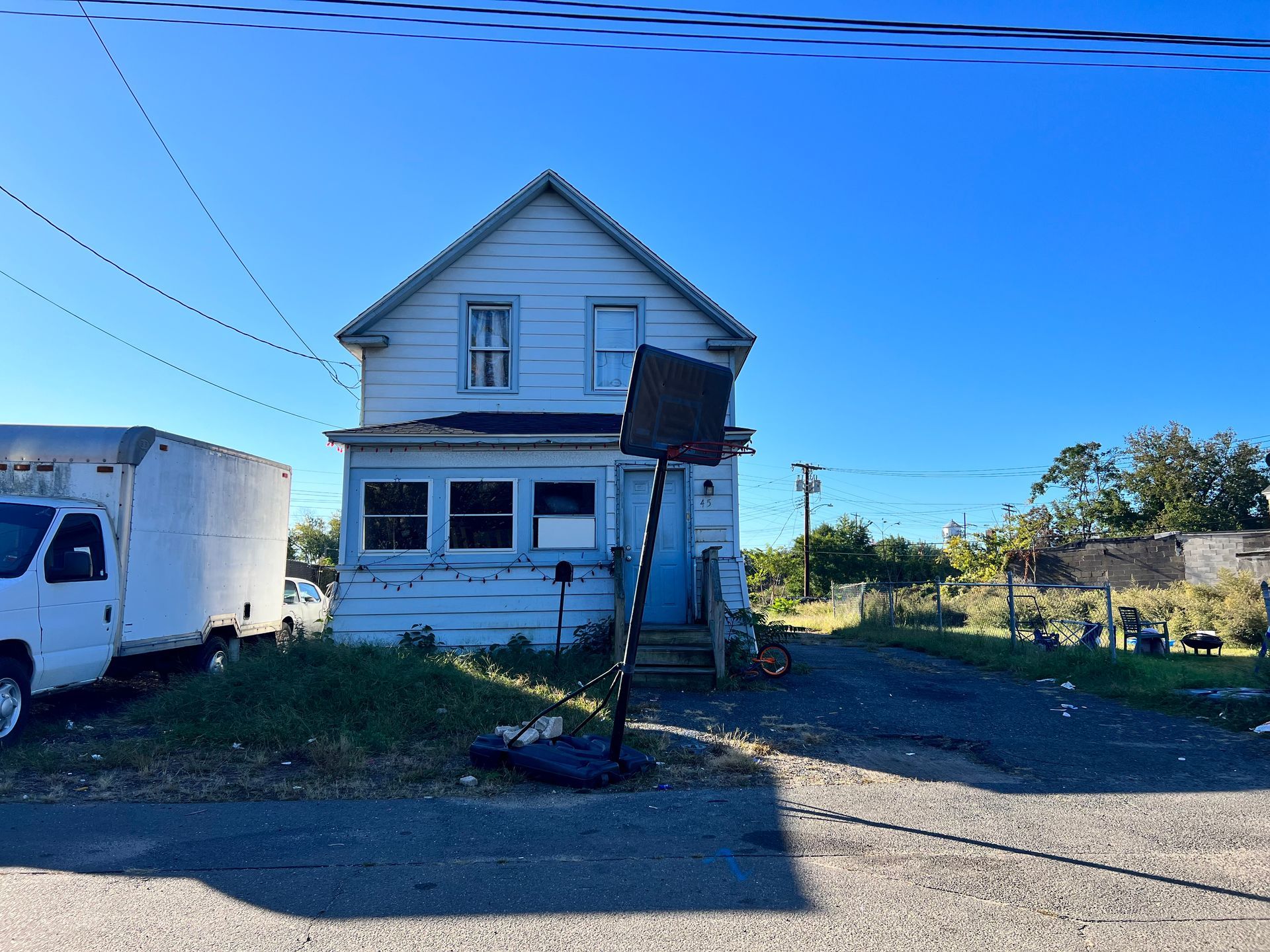 A dilapidated two-story house with a basketball hoop on a sunny day. A truck is parked to the left.