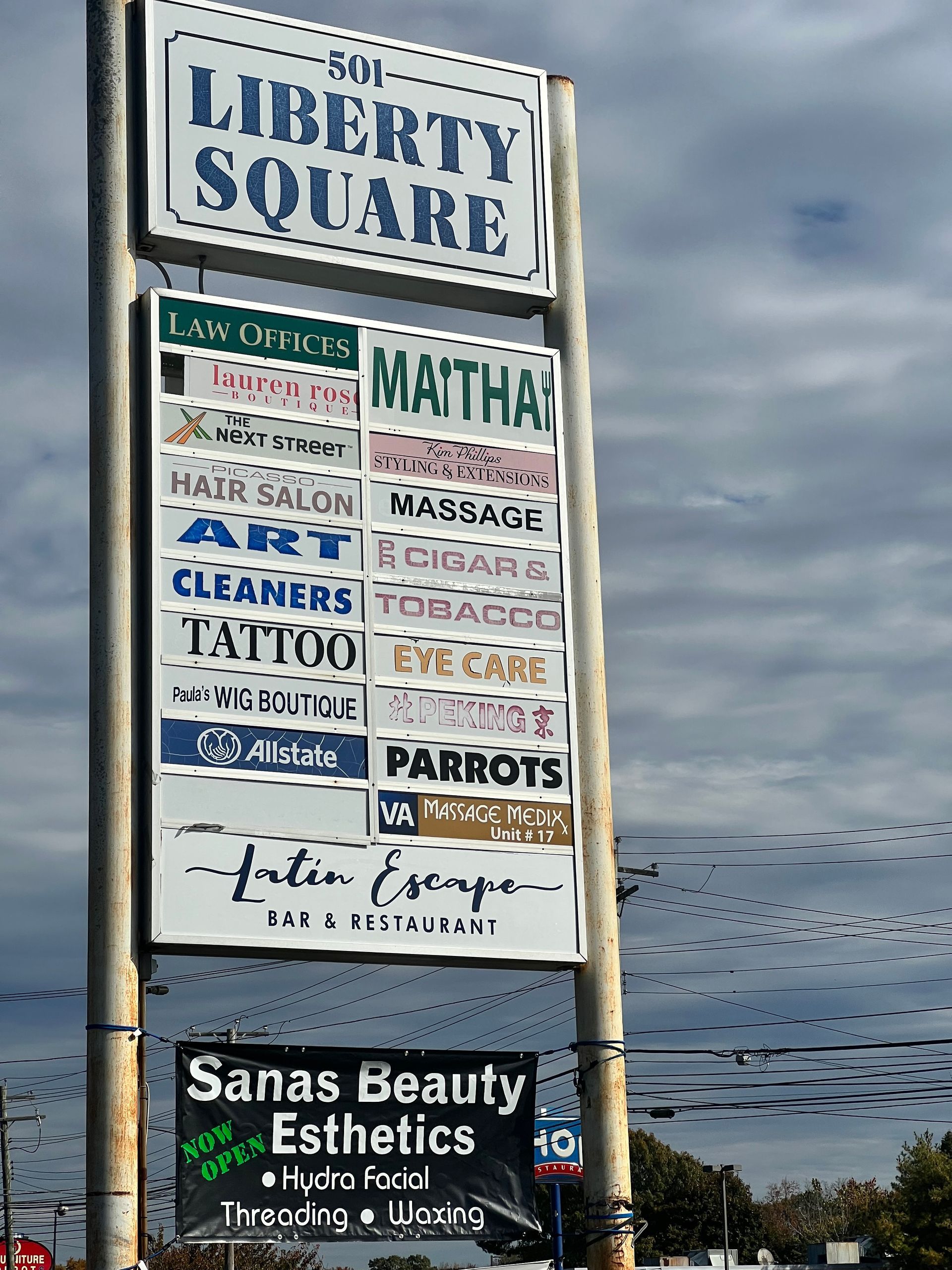 Sign for Liberty Square businesses. Includes names of shops like hair salons, cleaners, and eye care. Cloudy sky background.