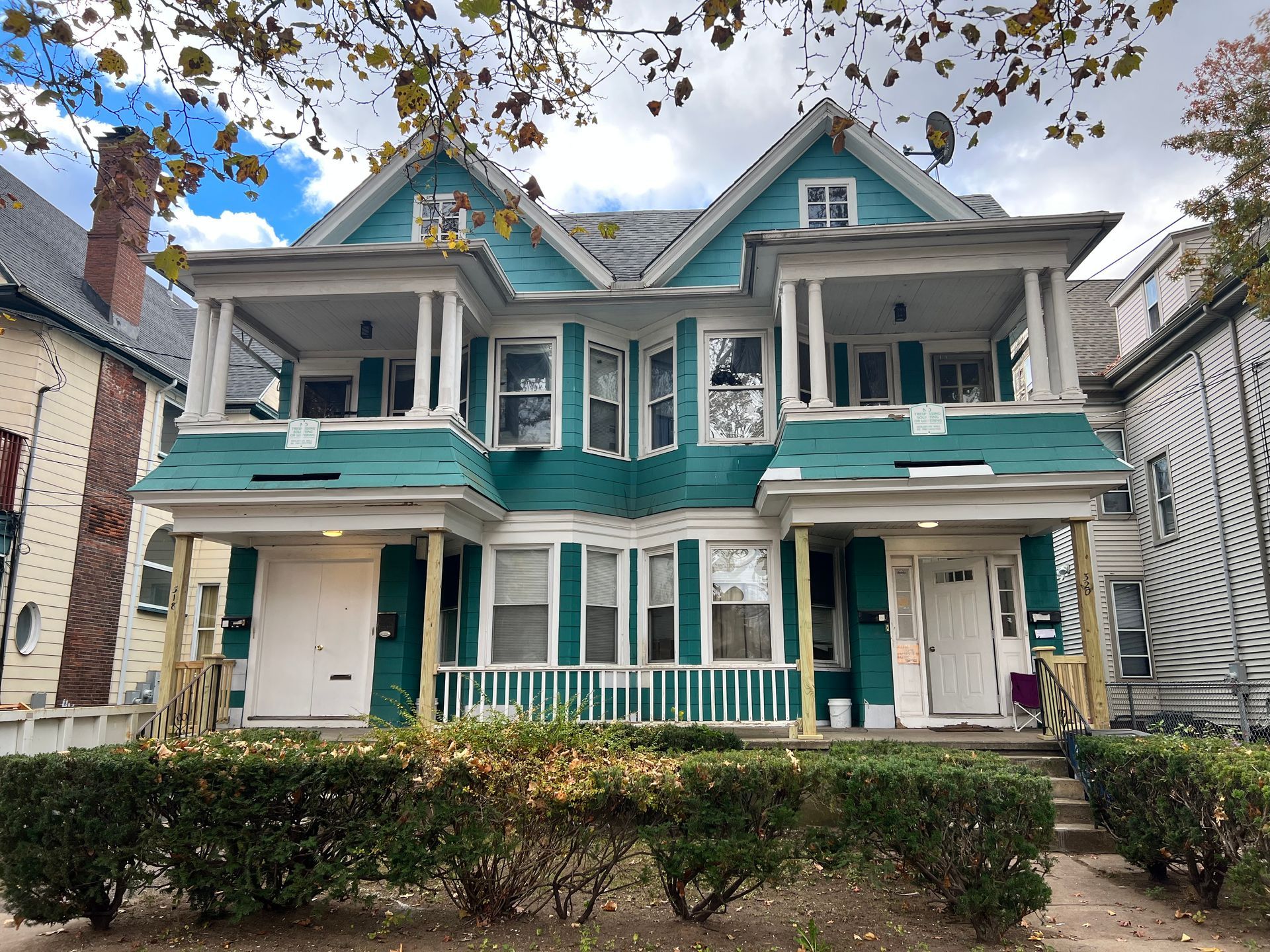 Two-story teal duplex with porches and white trim; green bushes line the front yard.